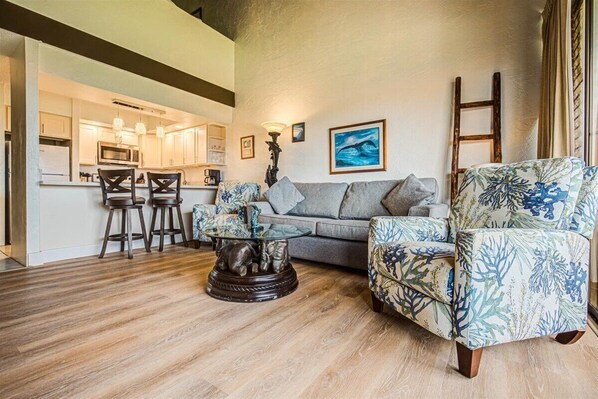 A modern living room with a grey sofa, patterned armchairs, a glass coffee table, and a kitchen in the background. - Spacious 3bd Papakea condo, the perfect getaway (Lahaina)