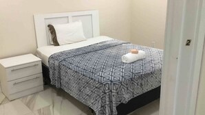 Un lit bien fait, recouvert d’une couvre-lit à motifs, avec un oreiller blanc et une serviette enroulée posée sur le lit. On y trouve une table de chevet blanche avec des tiroirs et une petite table de nuit avec une lampe.