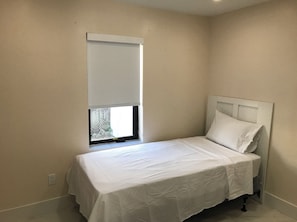 A small, single-bed room with a white bedspread, a white headboard, and a window with a white blind.