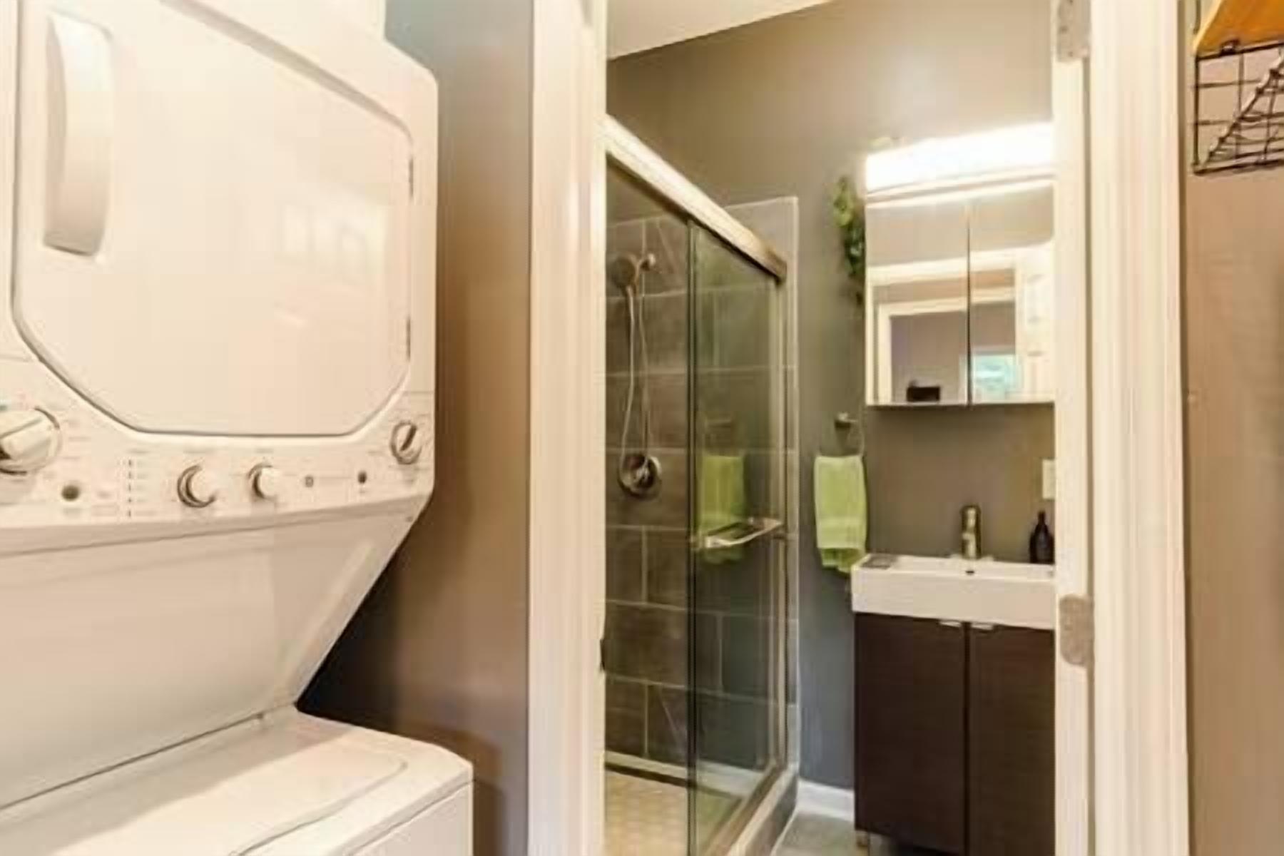 Un espace buanderie équipé d’un lave-linge et d’un sèche-linge, une cabine de douche en verre et une salle de bain avec un lavabo et un miroir.