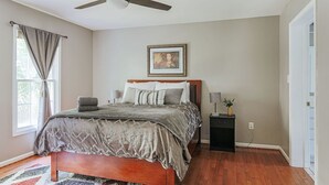 Un dormitorio con cama, ventilador de techo, mesita de noche con lámpara y un cuadro en la pared.