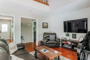 Een moderne woonkamer met een leren bank, een houten salontafel en een zwarte lederen fauteuil. Een flatscreen televisie hangt aan de muur, en er is een boekenkast met diverse spullen.