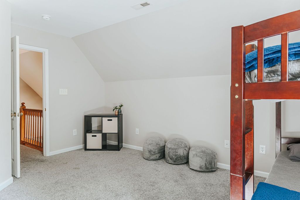 A room with a sloped ceiling, a wooden door with glass panels, a bed, a small table with a plant, and three round ottomans.