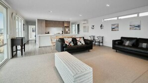 A spacious living area with a sofa, a coffee table, and a dining table. There is a kitchen in the background with wooden cabinets and a countertop.