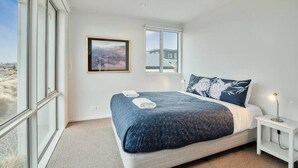A modern bedroom with a large bed, a small bedside table, a framed picture on the wall, and a view of a building through a window.