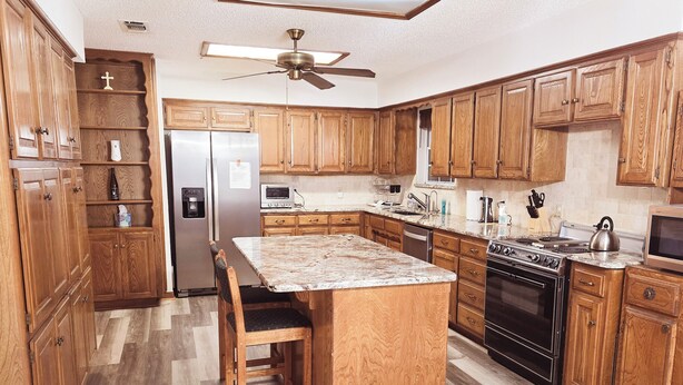 A kitchen with wooden cabinets, a central island, a refrigerator, a microwave, a stove, and a ceiling fan. - Country Living at its Best! (Roanoke)