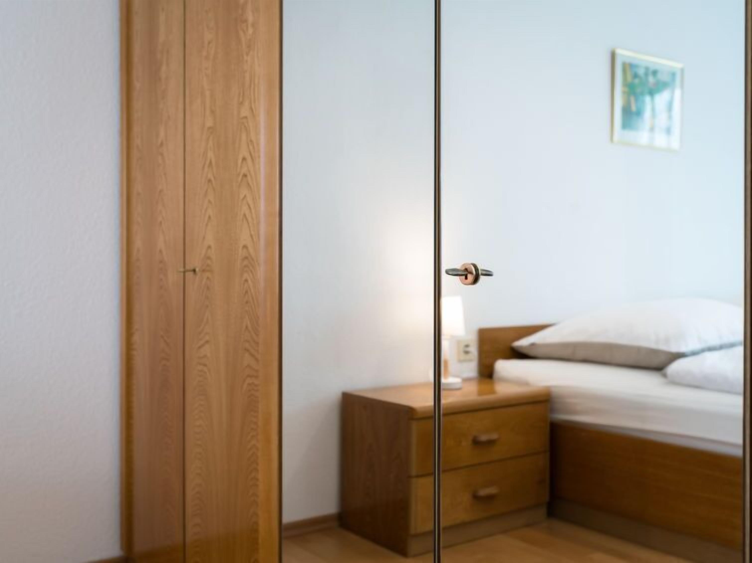 Un dormitorio con un armario de madera, una mesita de noche con lámpara y una cama con ropa de cama blanca.