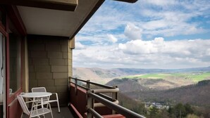 Un balcon avec une table et des chaises, donnant sur une vallée parsemée de collines et comprenant un village.