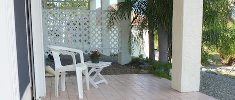 A covered patio area with a white plastic chair and table, surrounded by a white lattice fence, palm trees, and a gravel path.