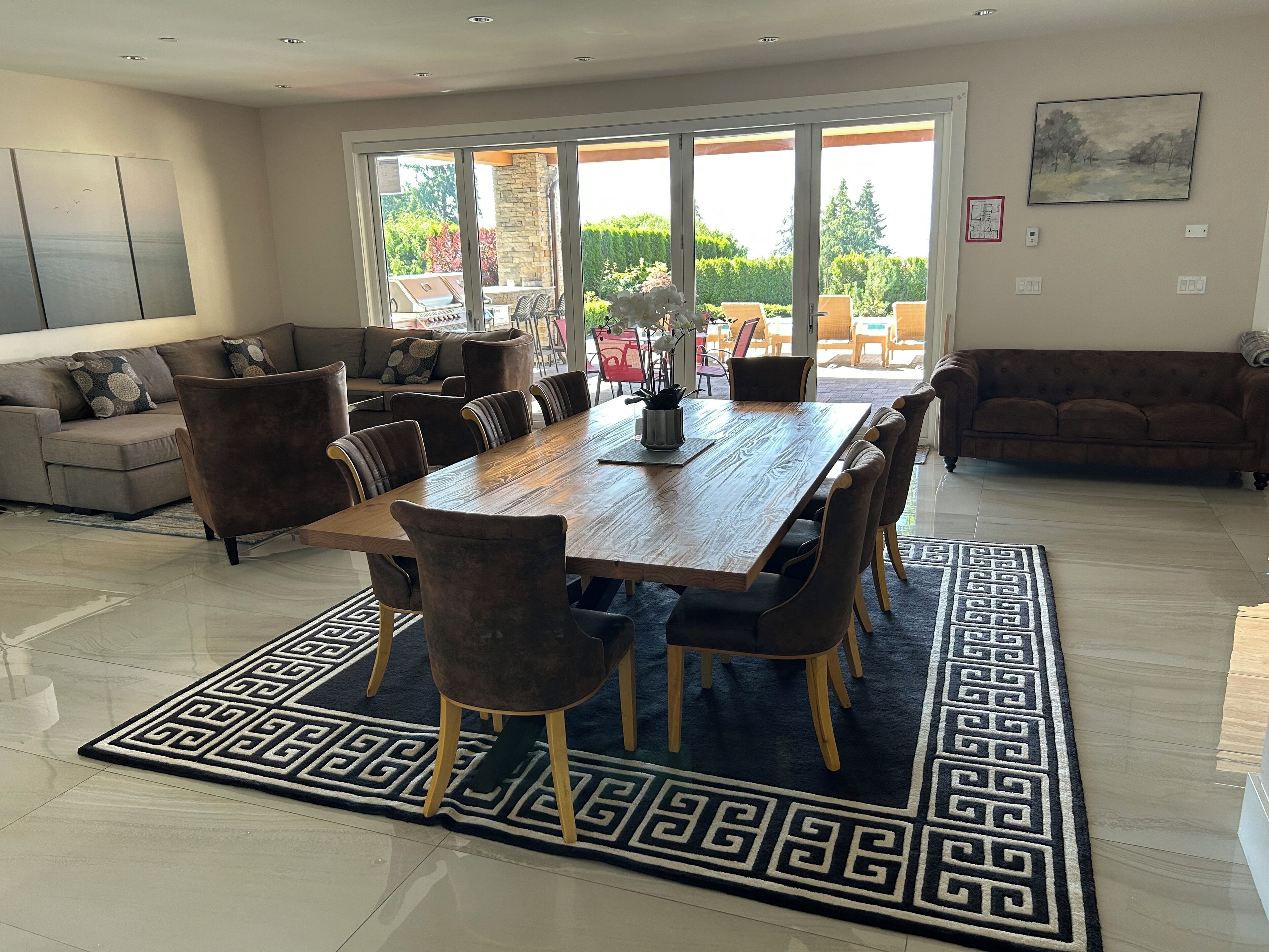 A spacious dining area with a large wooden table and matching chairs, a patterned rug, and a view of the outdoors through large sliding doors.