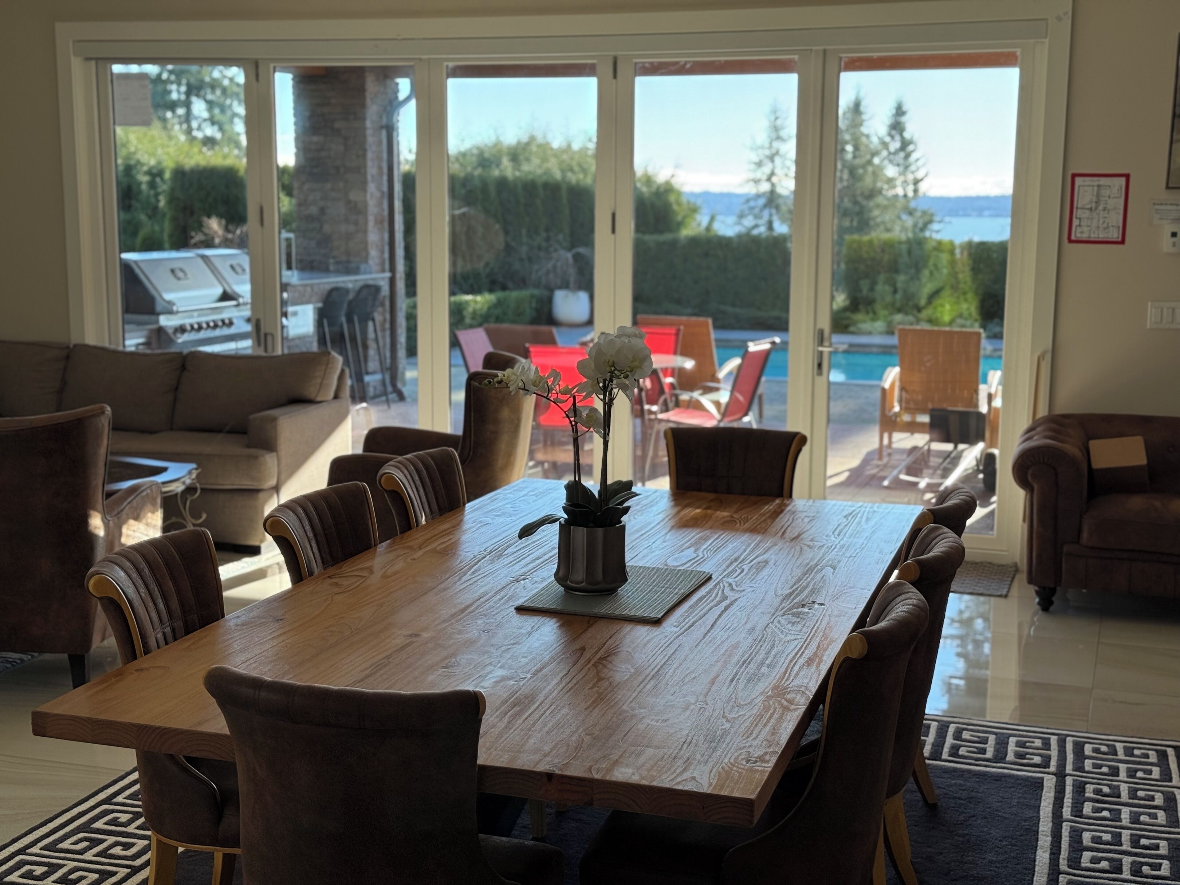 A spacious dining area with a wooden table and chairs, a potted orchid, and a view of a pool and greenery through large sliding glass doors.