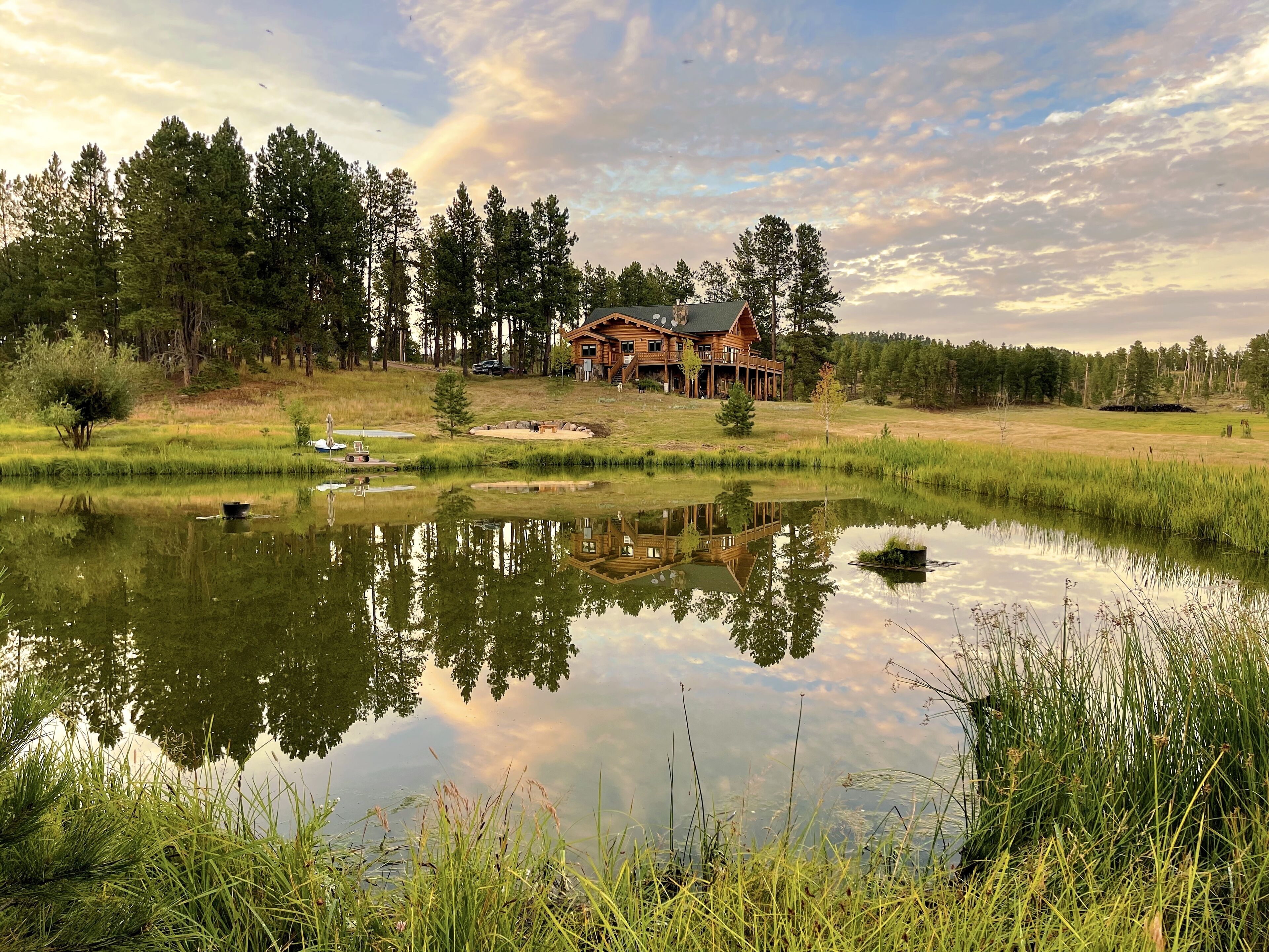 A large lodge with a prominent deck, surrounded by a pond and dense forest.
