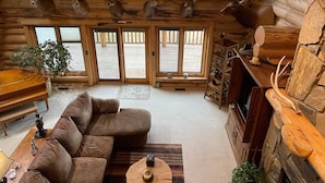 Een chaletinterieur met een ruime modulaire bank, een vleugelpiano en meerdere geweivleugels aan de wand bevestigd.