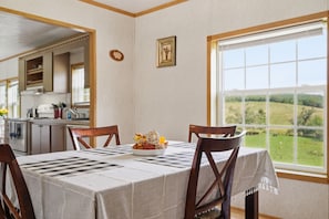 Ein Essbereich mit Tisch für vier Personen, Blick ins Freie durch ein großes Fenster und eine Küche im Hintergrund.