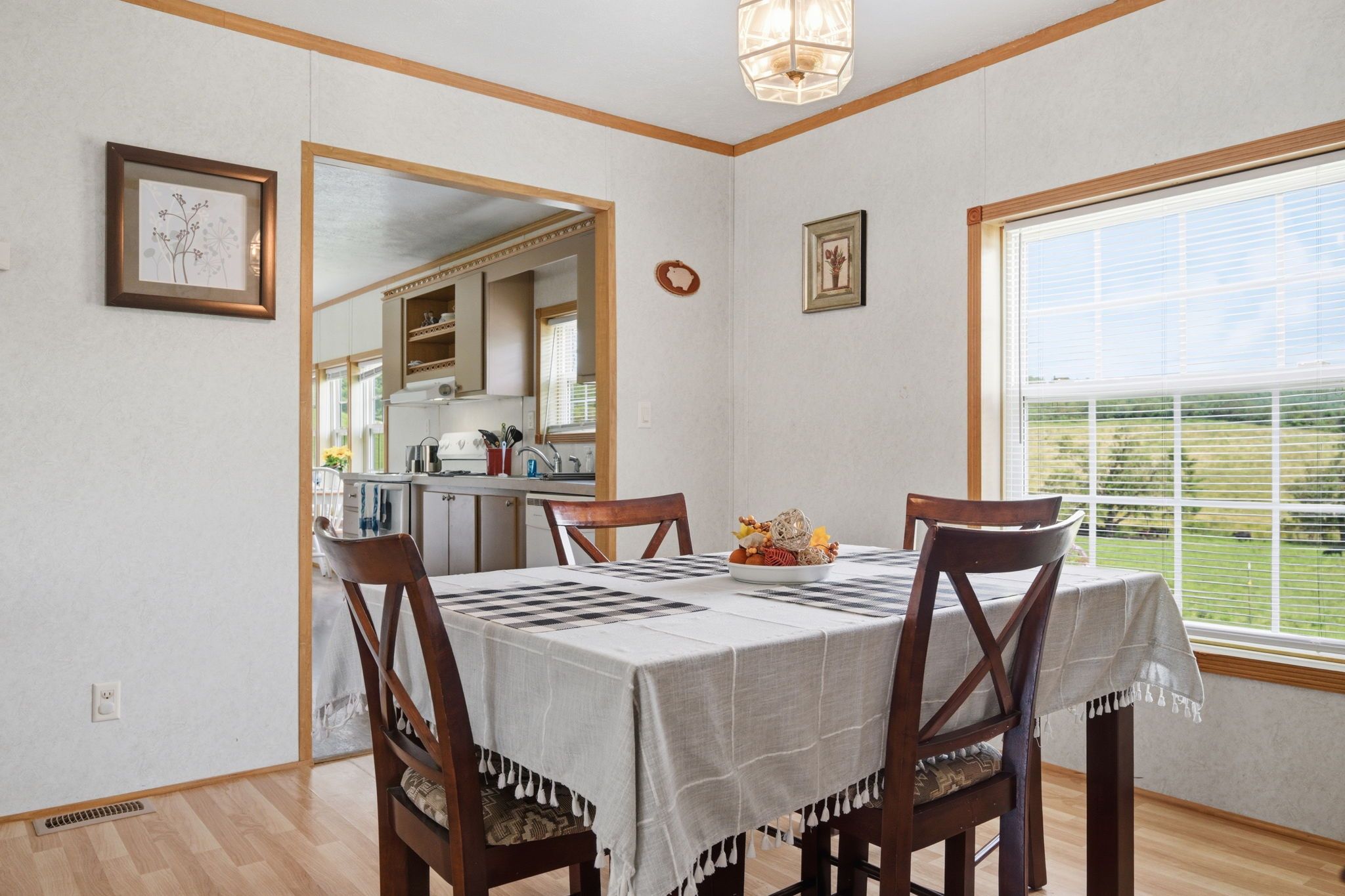 Une salle à manger avec une table dressée pour quatre personnes, des chaises en bois, une cuisine visible à travers une porte ouverte et une grande fenêtre avec des stores.
