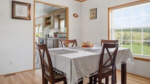 Un comedor con una mesa preparada para cuatro personas, sillas de madera, una cocina visible a través de una puerta abierta y un ventanal con persianas.