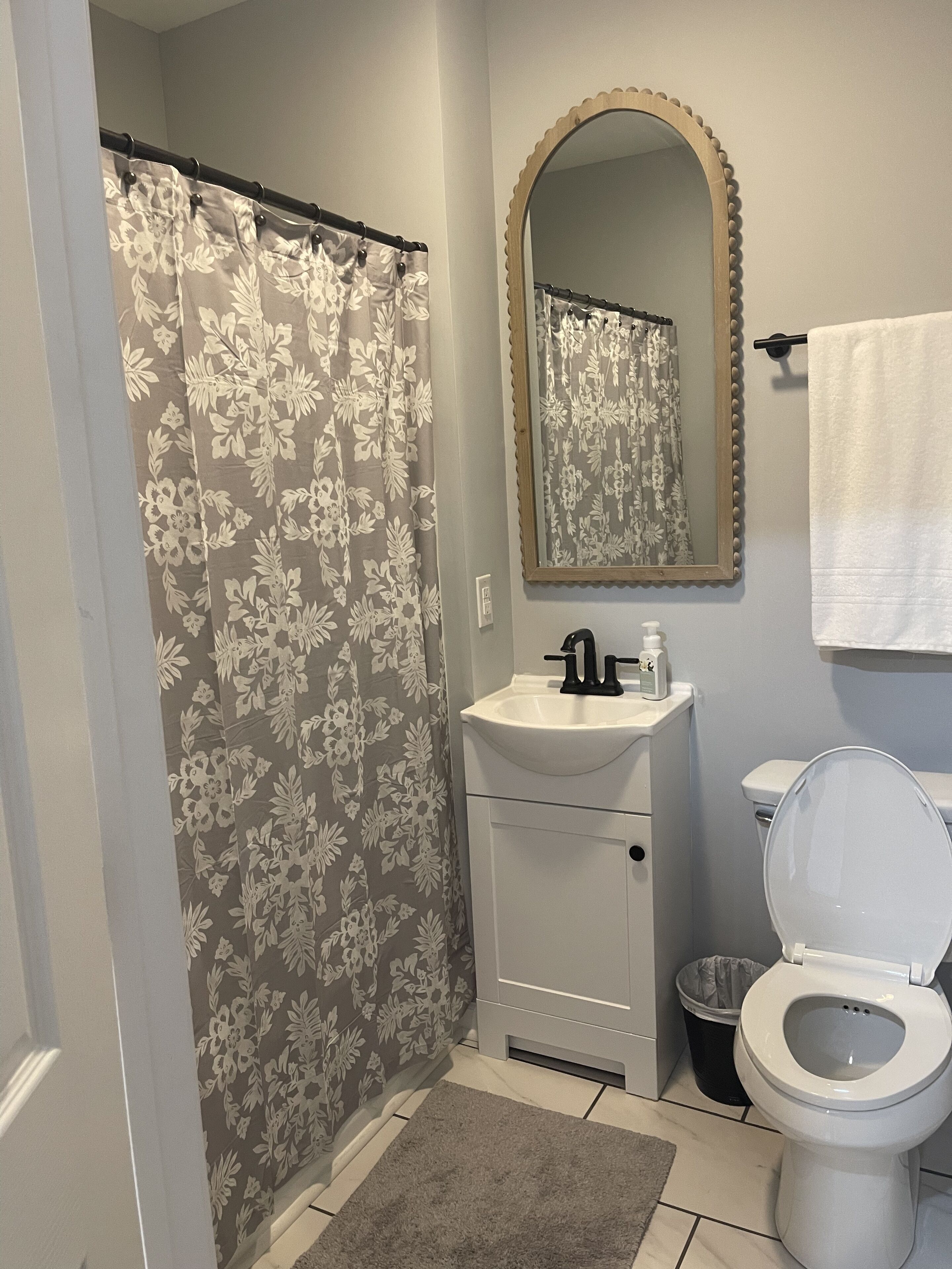 Une salle de bain équipée d’un rideau de douche, d’un lavabo, d’un miroir, de toilettes et d’une poubelle.