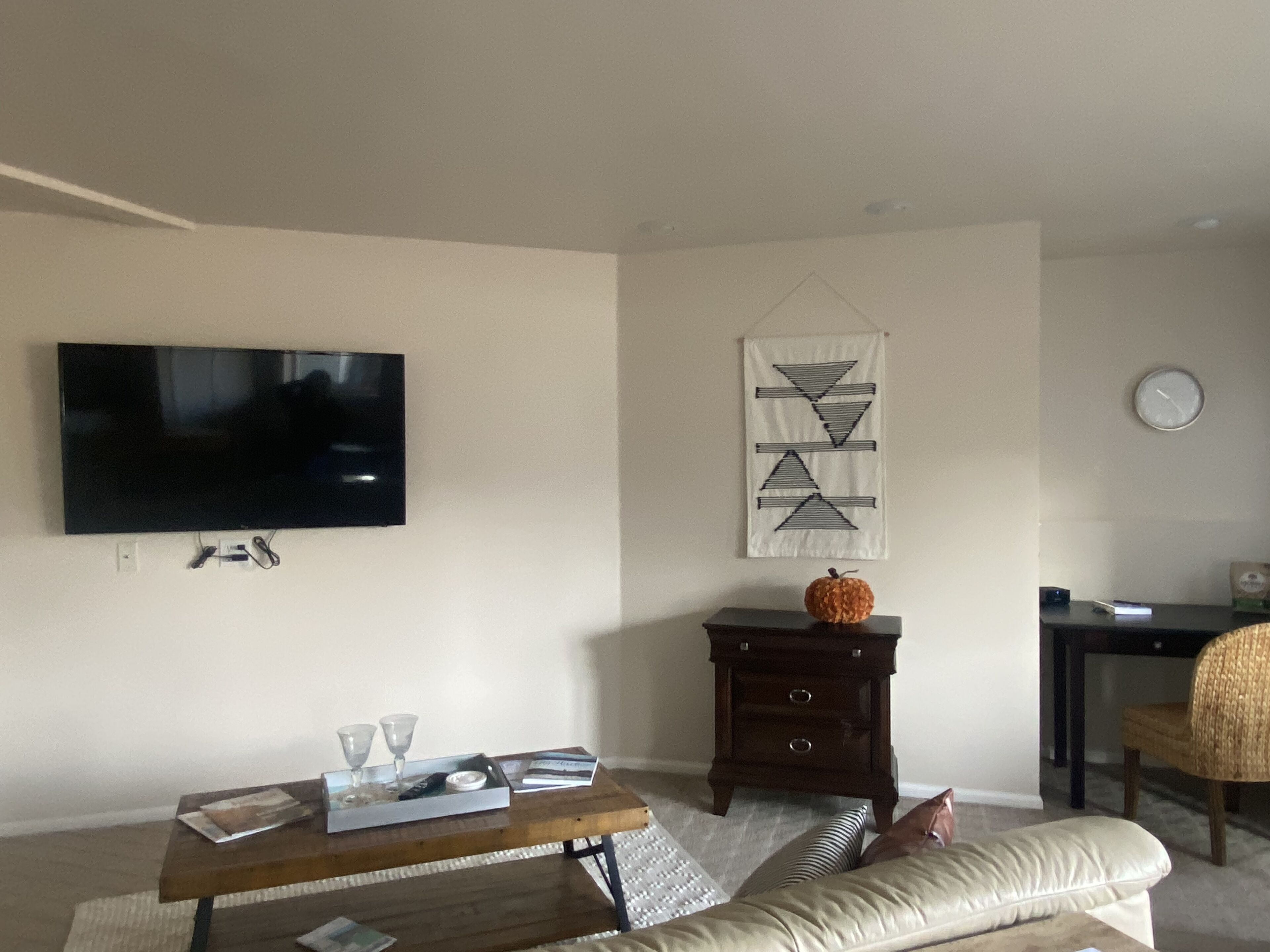 A living room with a flat-screen TV mounted on the wall, a small wooden table with a magazine and glasses, a decorative wall hanging, and a sofa with a cushion.