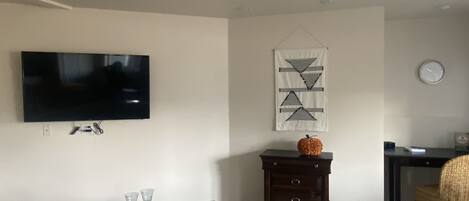 A living room with a flat-screen TV mounted on the wall, a small wooden table with a magazine and glasses, a decorative wall hanging, and a sofa with a cushion.