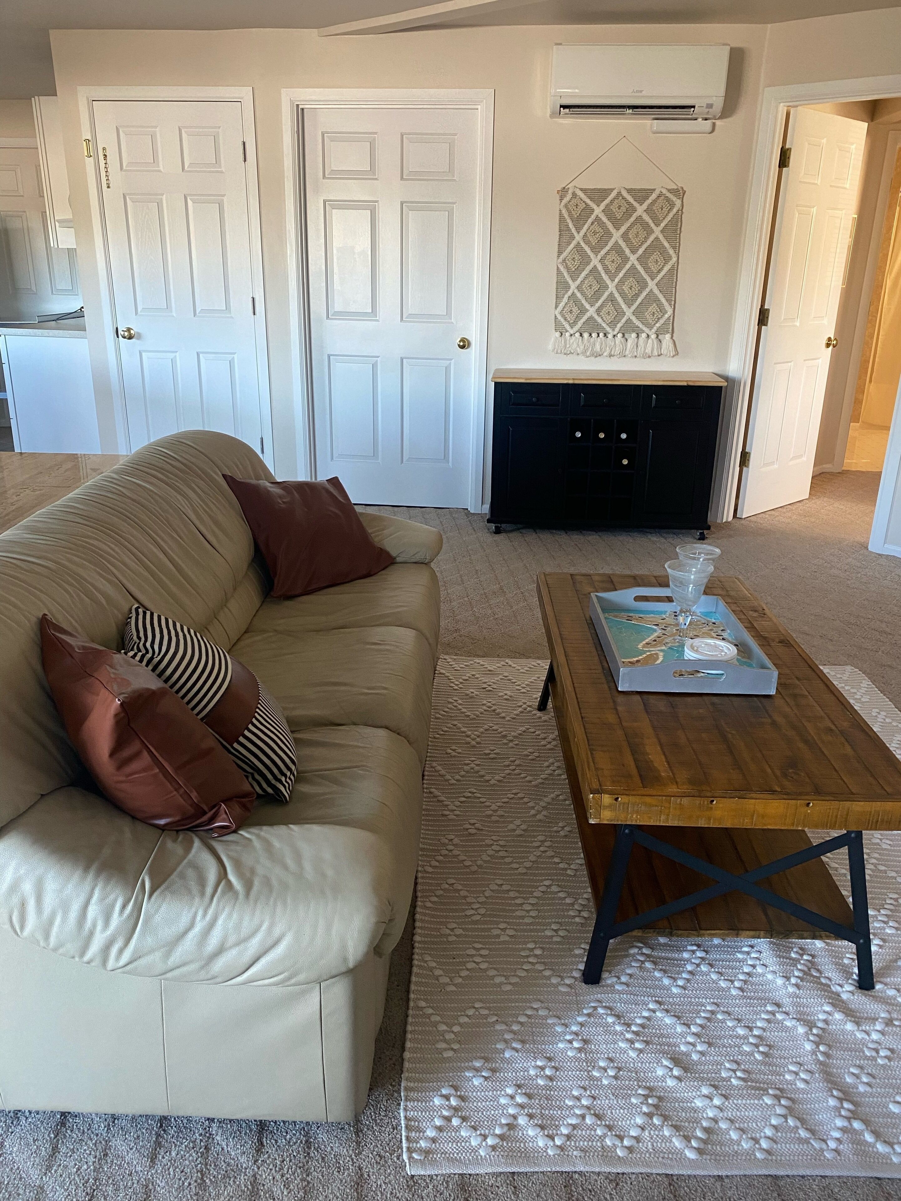 A living room with a beige sofa, a wooden coffee table, and a patterned rug. There are two white doors, a fireplace, and an air conditioning unit on the wall.