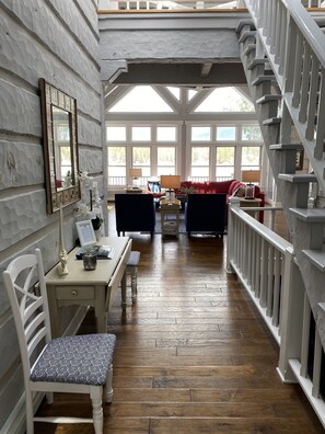 Relax in a spacious room with a high ceiling, featuring a desk and chair near a large window, a cozy seating area with a sofa and armchair, and a staircase leading to an upper level.