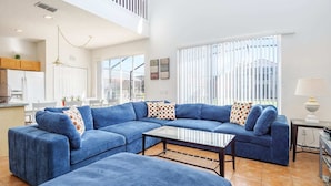 Un salón amplio con un sofá seccional azul grande, una mesa de centro de vidrio y vistas al exterior a través de grandes ventanales.
