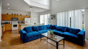 Amplio salón con un sofá seccional azul grande, una mesa de centro de vidrio y una zona de cocina con armarios de madera y electrodomésticos modernos.