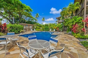 A pool area with lounge chairs and tables, surrounded by tropical plants and a clear blue sky.
