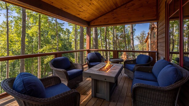 A covered deck with wicker furniture and a central fire feature, surrounded by tall trees. - MountainAire at Eagles Nest -  Views, firepit, gas fire table, Eagles Nest amenities! (Banner Elk)