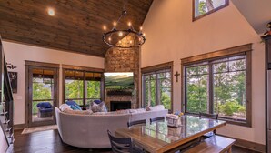 A spacious living area with a stone fireplace, a large window with a view of greenery, and a wooden ceiling. - MountainAire at Eagles Nest -  Views, firepit, gas fire table, Eagles Nest amenities! (Banner Elk)
