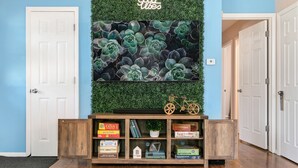 Un pasillo con una consola de madera que muestra libros y juegos, una pared verde con plantas suculentas y una puerta a la izquierda.