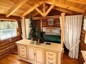 Interior de cabana com teto de madeira, cama com colcha verde, cómoda com gavetas e janela com persianas.