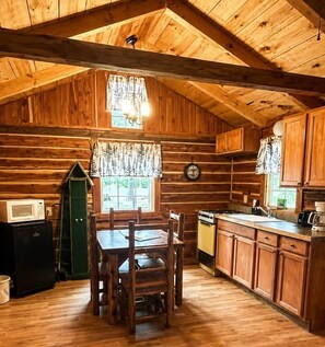 Interior rústico de uma cabana de madeira, com mesa de jantar, cadeiras, área de cozinha com armários e uma janela com cortinas.