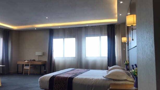 A hotel room with a bed, desk, and chair, large windows, and a lamp.