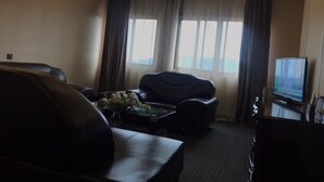 A hotel room with a sofa, a coffee table with flowers, a TV, and a view of the city through large windows.