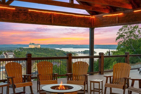 A covered outdoor seating area with wooden chairs and a central fire pit, overlooking a scenic view of a lake and distant buildings.