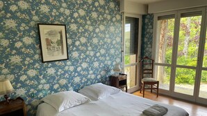 Une chambre avec un grand lit, un papier peint à motifs floraux, un tableau encadré au mur, une chaise et une fenêtre donnant sur un paysage verdoyant.