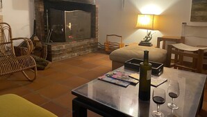 Un salon chaleureux avec une cheminée, un canapé confortable, une table à manger avec une bouteille de vin et des verres, et une lampe.