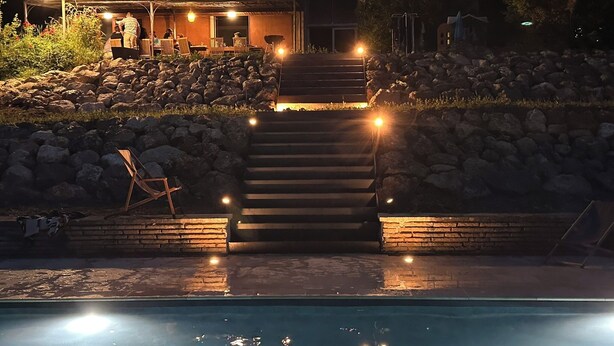 Un espace piscine avec des marches menant à un coin salon extérieur abrité, entouré de rochers et d’un mur de briques.