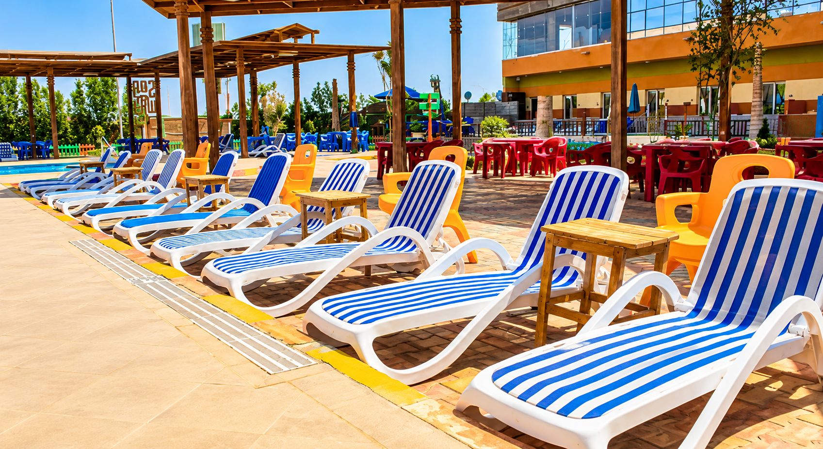 Un espace aménagé au bord de la piscine, avec des chaises longues et des tables, entouré d’un bâtiment doté de grandes fenêtres.