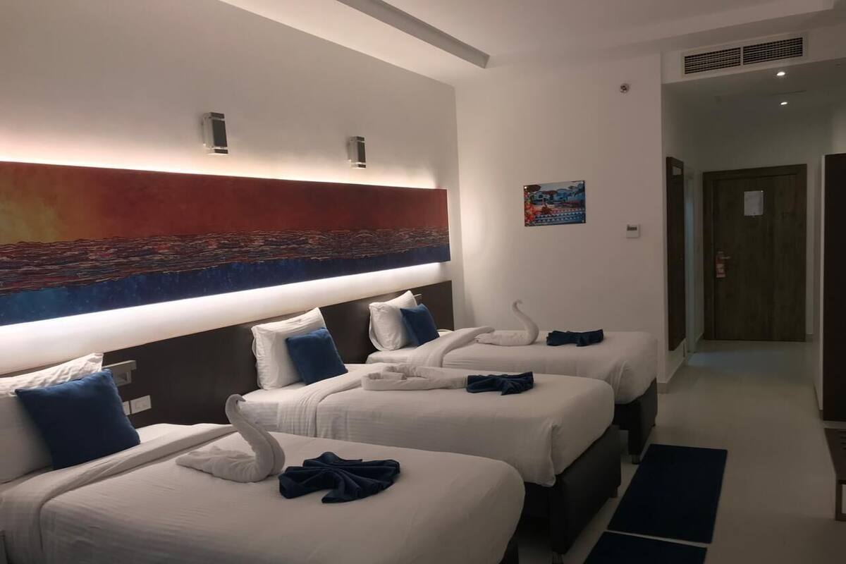 Ein Hotelzimmer mit drei Betten, jedes mit weißer Bettwäsche und blauen Kissen. Darüber hängt ein großes, abstraktes Gemälde an der Wand.