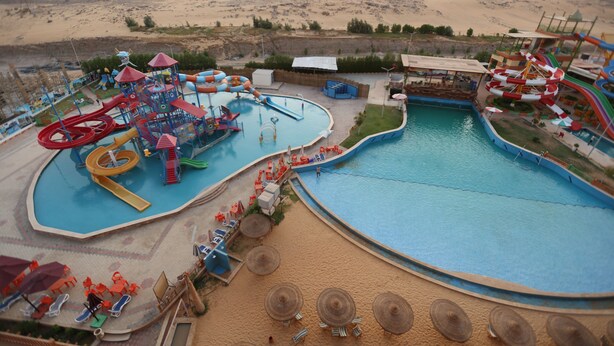 A water park with multiple slides and pools, surrounded by a sandy area and buildings in the background. - Gloria Aqua Park Hotel (Aswan)