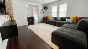 A modern living room with a dark wood floor, a large sectional sofa with colorful cushions, a white shag rug, and a flat-screen TV mounted on the wall. - Large, modern 2 bedroom in the heart of Forest Park (Forest Park)