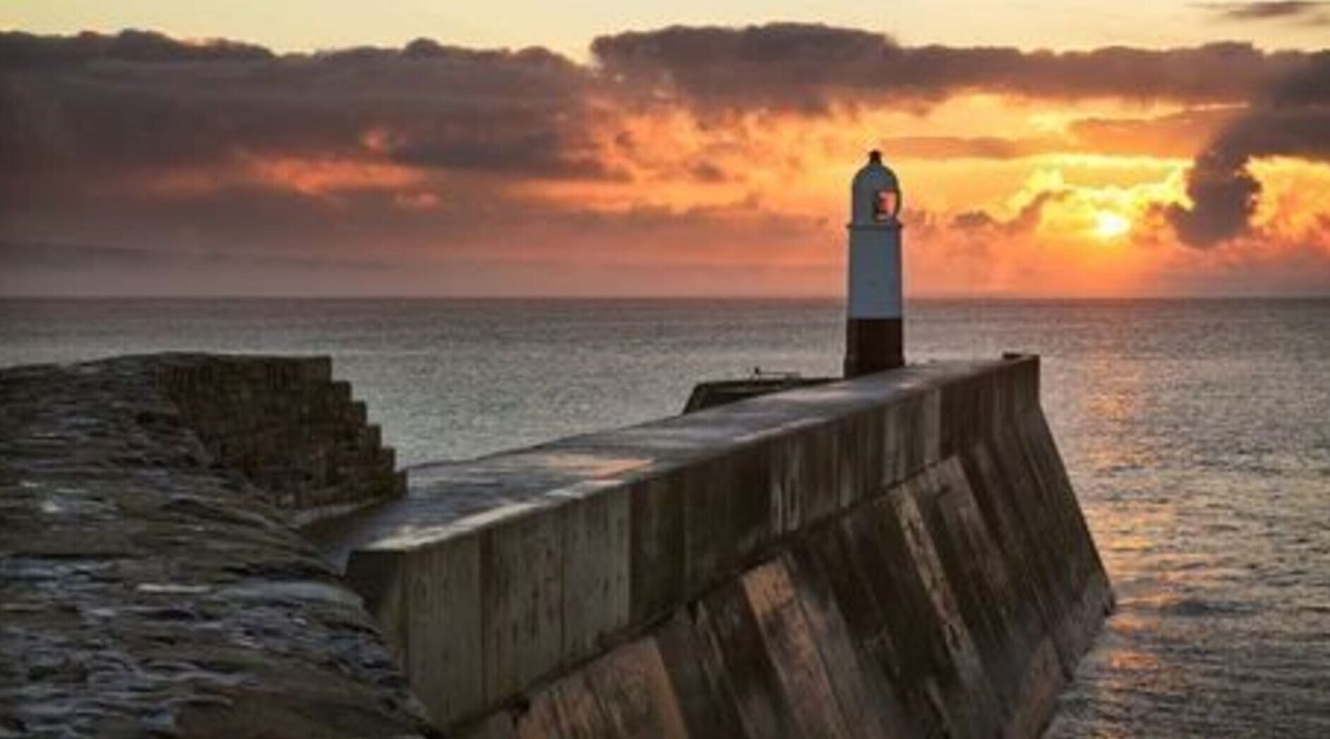 A lighthouse on a concrete pier with a sunset in the background.