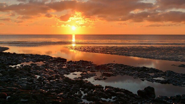 Un coucher de soleil sur une côte rocheuse, avec une eau calme qui reflète le ciel.