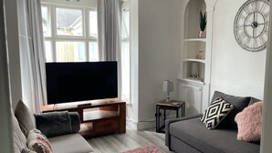 Un salon moderne avec un téléviseur à écran plat, des canapés gris, un tapis rose et une horloge décorative accrochée au mur.