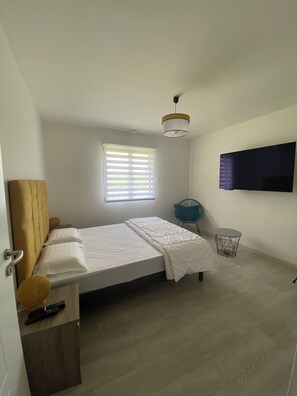 Een moderne slaapkamer met een bed, een aan de muur bevestigde televisie, een raam met zonwering en een klein rond tafeltje.
