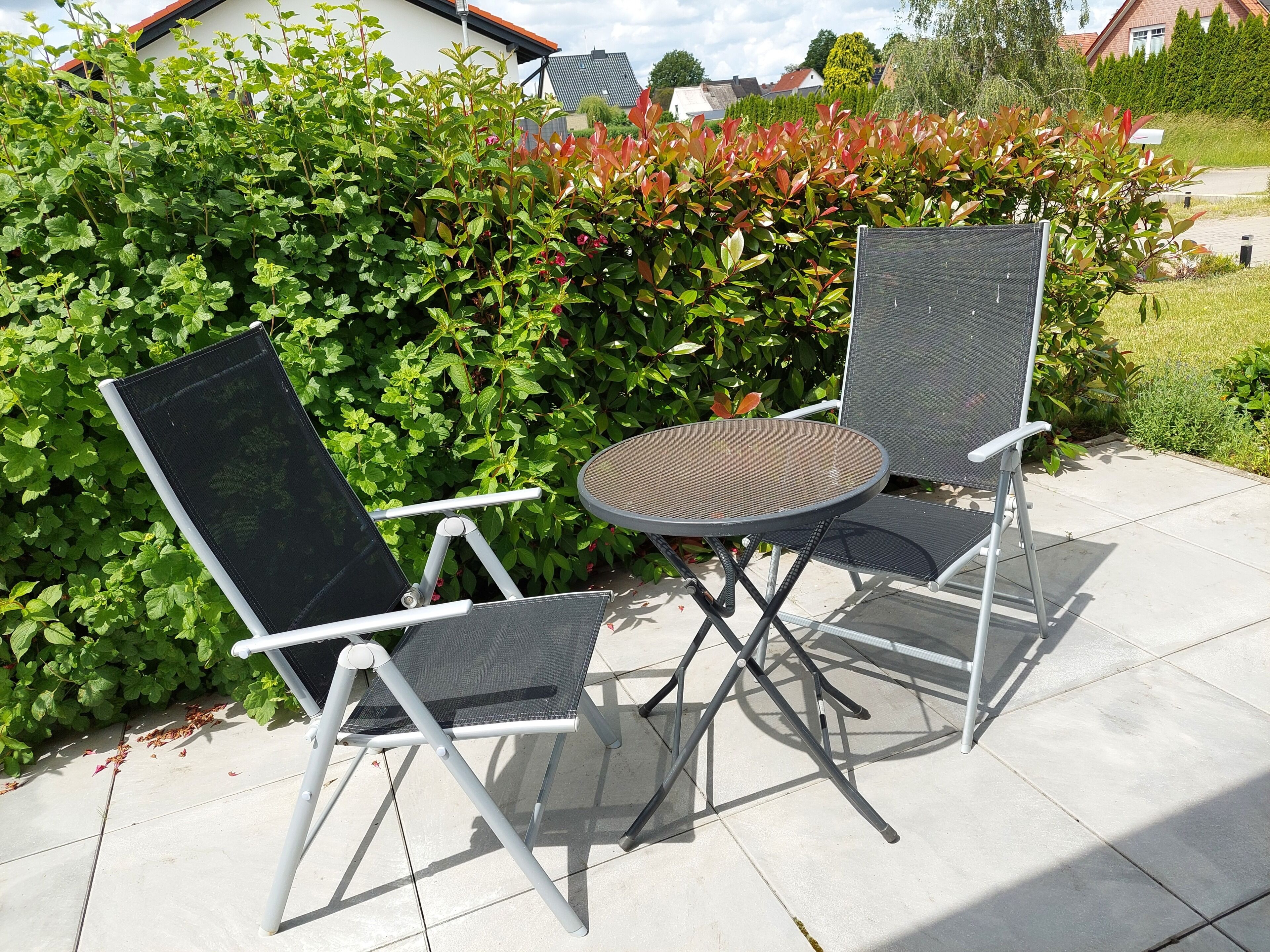 Ein Außenbereich mit Sitzgelegenheit, zwei Stühlen und einem Tisch, umgeben von Pflanzen und einer Hecke.