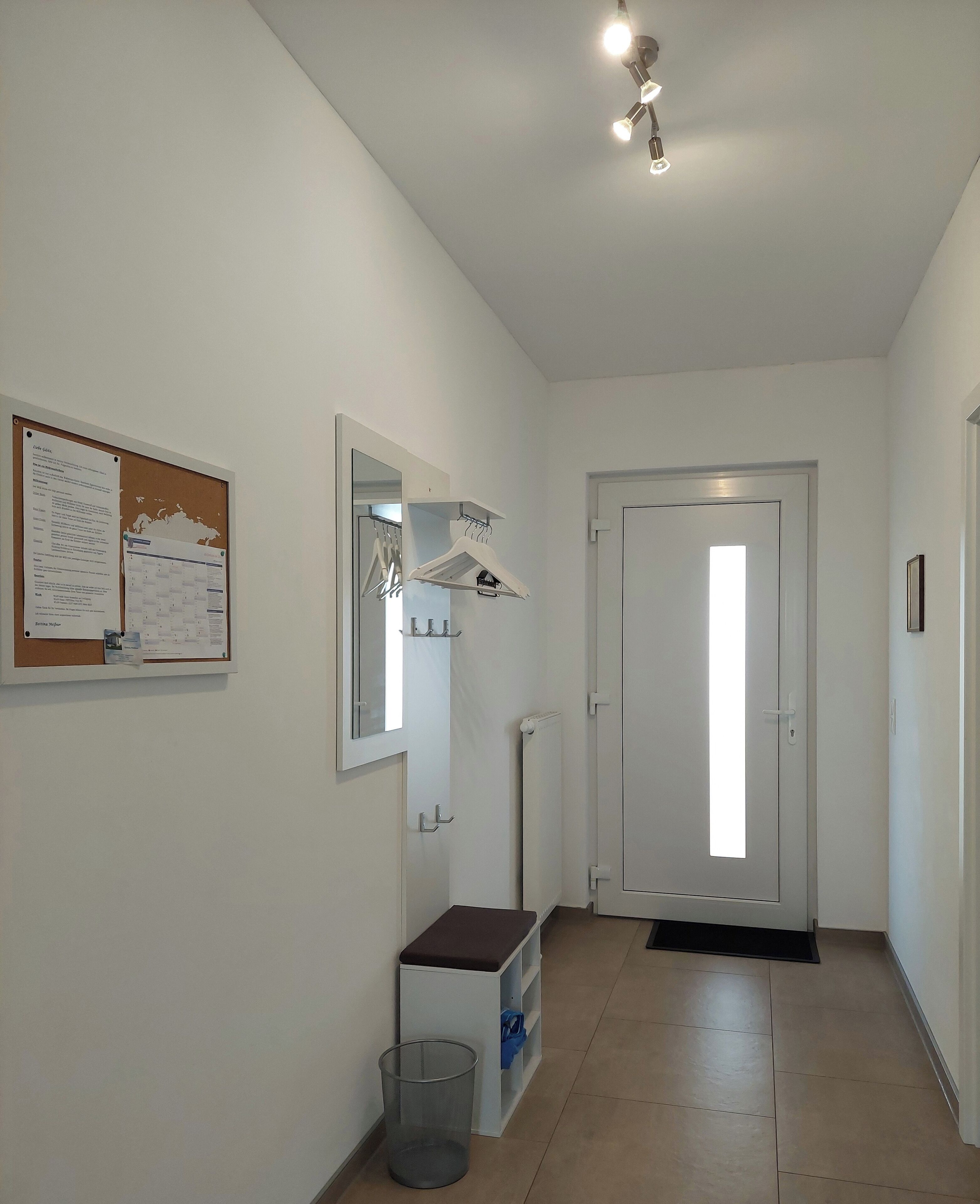Un couloir avec un tableau d’affichage, un porte-manteau, une porte et un radiateur.