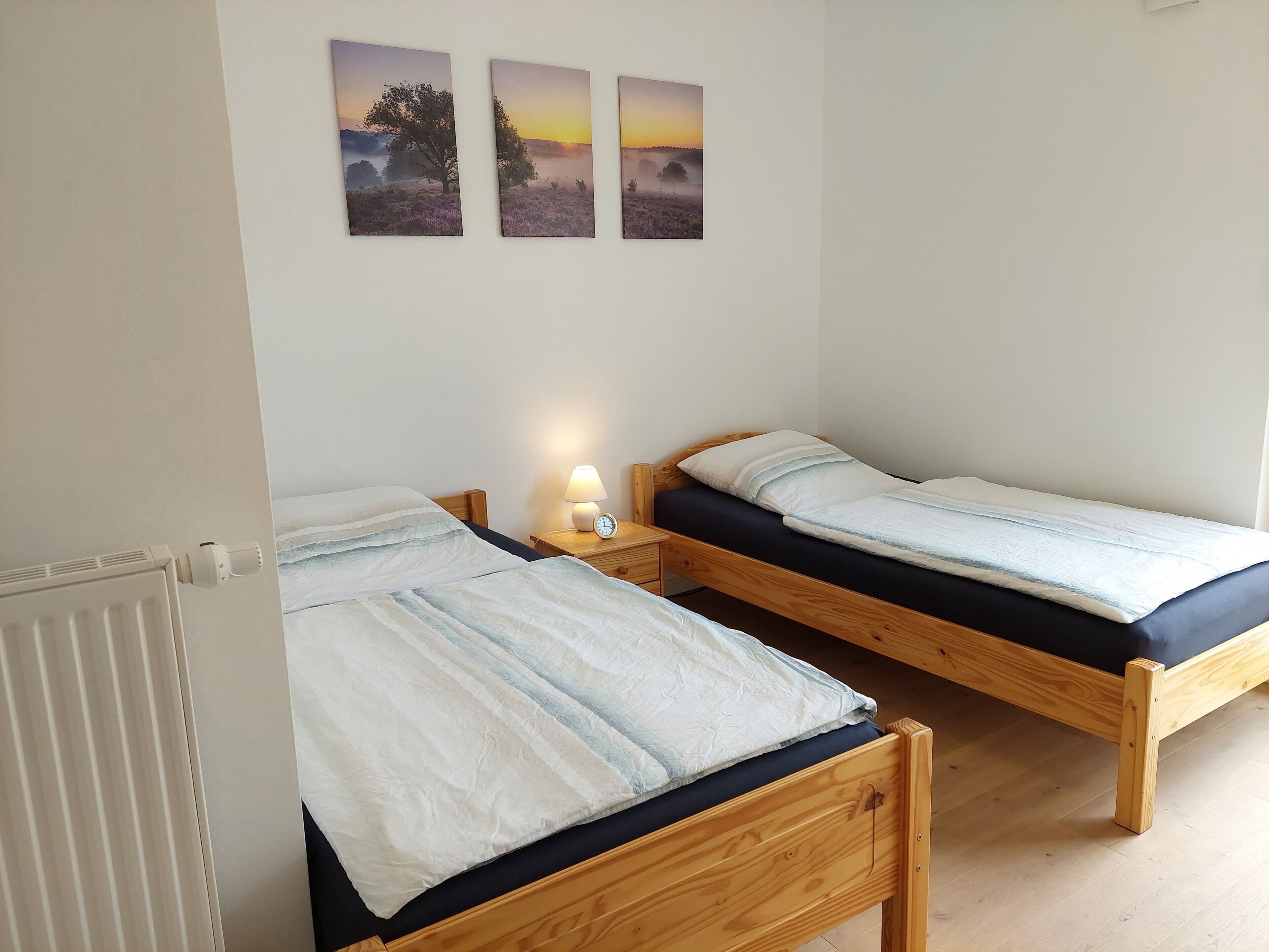 Une chambre avec deux lits en bois, une table de chevet avec une lampe et trois tableaux représentant des paysages accrochés au mur.